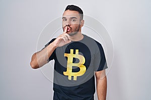 Young hispanic man wearing bitcoin t shirt asking to be quiet with finger on lips