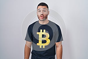 Young hispanic man wearing bitcoin t shirt making fish face with lips, crazy and comical gesture