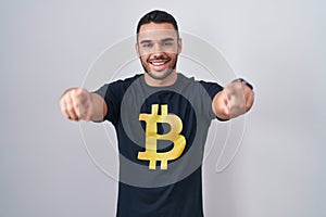 Young hispanic man wearing bitcoin t shirt pointing to you and the camera with fingers, smiling positive and cheerful