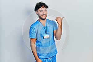 Young hispanic man wearing blue male nurse uniform smiling with happy face looking and pointing to the side with thumb up