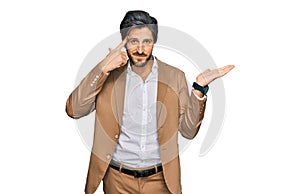 Young hispanic man wearing business clothes confused and annoyed with open palm showing copy space and pointing finger to forehead