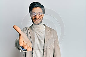 Young hispanic man wearing business jacket and glasses smiling friendly offering handshake as greeting and welcoming