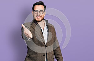 Young hispanic man wearing business jacket and glasses smiling friendly offering handshake as greeting and welcoming