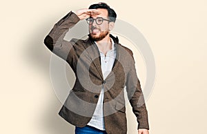 Young hispanic man wearing business jacket and glasses very happy and smiling looking far away with hand over head