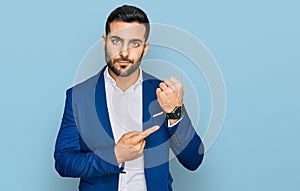 Young hispanic man wearing business jacket in hurry pointing to watch time, impatience, looking at the camera with relaxed