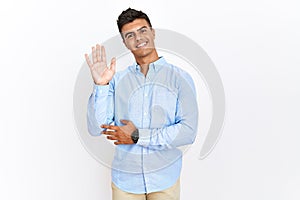 Young hispanic man wearing business shirt standing over isolated background waiving saying hello happy and smiling, friendly