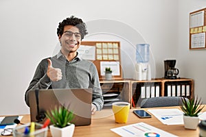 Young hispanic man wearing business style sitting on desk at office doing happy thumbs up gesture with hand