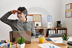 Young hispanic man wearing business style sitting on desk at office very happy and smiling looking far away with hand over head