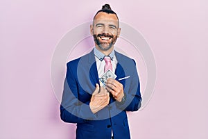 Young hispanic man wearing business suit holding dollar smiling and laughing hard out loud because funny crazy joke