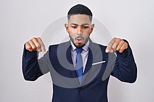 Young hispanic man wearing business suit and tie pointing down with fingers showing advertisement, surprised face and open mouth
