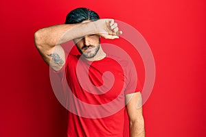 Young hispanic man wearing casual red t shirt covering eyes with arm, looking serious and sad