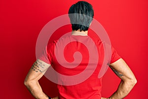 Young hispanic man wearing casual red t shirt standing backwards looking away with arms on body