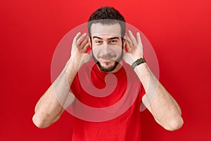 Young hispanic man wearing casual red t shirt trying to hear both hands on ear gesture, curious for gossip