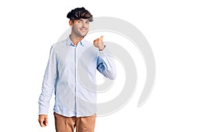 Young hispanic man wearing casual shirt smiling with happy face looking and pointing to the side with thumb up