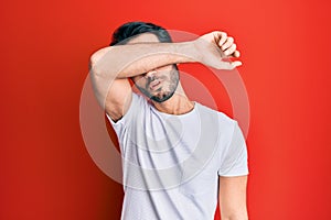Young hispanic man wearing casual white tshirt covering eyes with arm, looking serious and sad