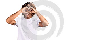 Young hispanic man wearing casual white tshirt doing heart shape with hand and fingers smiling looking through sign