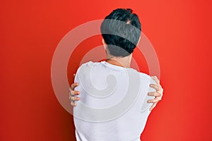 Young hispanic man wearing casual white tshirt hugging oneself happy and positive from backwards