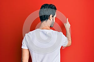 Young hispanic man wearing casual white tshirt posing backwards pointing ahead with finger hand