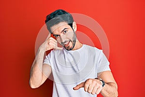 Young hispanic man wearing casual white tshirt smiling doing talking on the telephone gesture and pointing to you