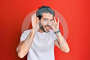 Young hispanic man wearing casual white tshirt trying to hear both hands on ear gesture, curious for gossip