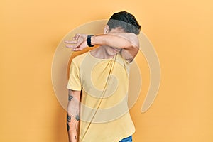 Young hispanic man wearing casual yellow t shirt covering eyes with arm, looking serious and sad