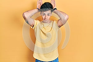 Young hispanic man wearing casual yellow t shirt doing bunny ears gesture with hands palms looking cynical and skeptical