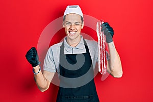 Young hispanic man wearing cook apron and holding meat screaming proud, celebrating victory and success very excited with raised