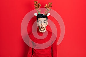 Young hispanic man wearing cute christmas reindeer horns making fish face with lips, crazy and comical gesture