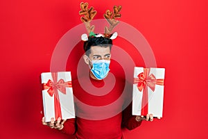 Young hispanic man wearing cute christmas reindeer horns and medical mask holding gifts angry and mad screaming frustrated and