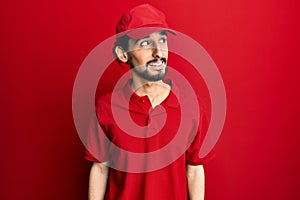 Young hispanic man wearing delivery uniform and cap looking away to side with smile on face, natural expression