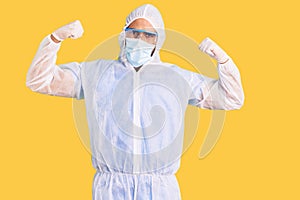 Young hispanic man wearing doctor protection coronavirus uniform and medical mask showing arms muscles smiling proud