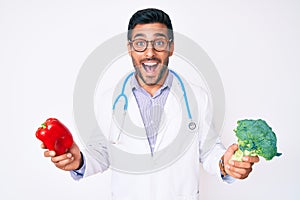 Young hispanic man wearing doctor stethoscope holding red pepper and broccoli smiling and laughing hard out loud because funny