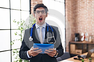 Young hispanic man wearing doctor stethoscope at the office angry and mad screaming frustrated and furious, shouting with anger