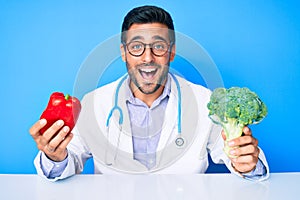Young hispanic man wearing doctor uniform holding red pepper and broccoli smiling and laughing hard out loud because funny crazy