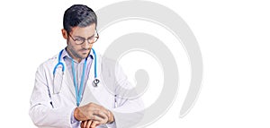 Young hispanic man wearing doctor uniform and stethoscope checking the time on wrist watch, relaxed and confident