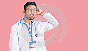 Young hispanic man wearing doctor uniform and stethoscope confuse and wondering about question