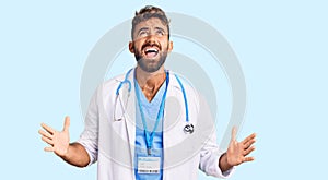 Young hispanic man wearing doctor uniform and stethoscope crazy and mad shouting and yelling with aggressive expression and arms