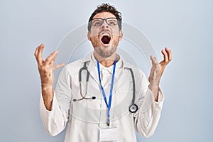 Young hispanic man wearing doctor uniform and stethoscope crazy and mad shouting and yelling with aggressive expression and arms