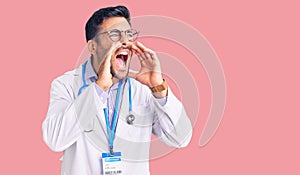 Young hispanic man wearing doctor uniform and stethoscope shouting angry out loud with hands over mouth