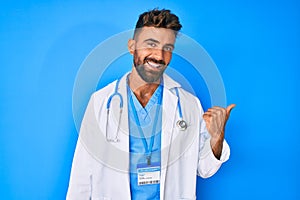 Young hispanic man wearing doctor uniform and stethoscope smiling with happy face looking and pointing to the side with thumb up