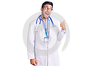 Young hispanic man wearing doctor uniform and stethoscope smiling with happy face looking and pointing to the side with thumb up