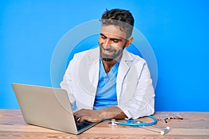 Young hispanic man wearing doctor uniform working at the clinic looking positive and happy standing and smiling with a confident