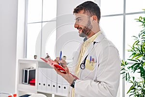 Young hispanic man wearing doctor uniform writing on clipboard at clinic