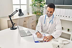 Young hispanic man wearing doctor uniform writing on clipboard at clinic