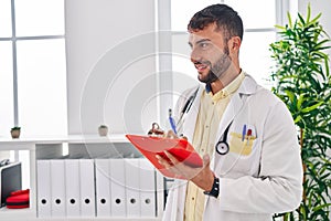 Young hispanic man wearing doctor uniform writing on clipboard at clinic