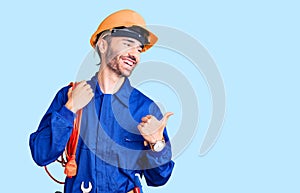 Young hispanic man wearing elecrician uniform holding cable pointing thumb up to the side smiling happy with open mouth