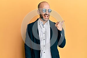 Young hispanic man wearing fashion sunglasses and business jacket pointing thumb up to the side smiling happy with open mouth