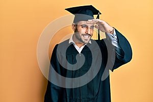 Young hispanic man wearing graduation cap and ceremony robe very happy and smiling looking far away with hand over head