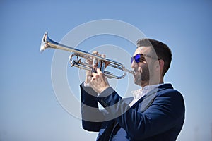 Young Hispanic man, wearing a jacket and sunglasses, playing a pretty, silvery trumpet outdoors. Concept, music, instruments,
