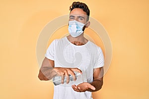 Young hispanic man wearing medical mask holding hand sanitizer gel looking at the camera blowing a kiss being lovely and sexy